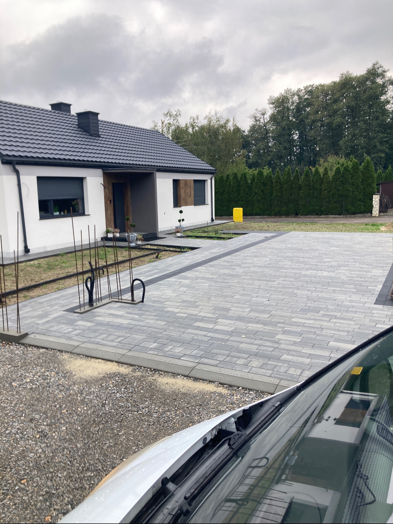 Nowoczesny podjazd z szarej kostki brukowej przed domem z białymi ścianami i ciemną stolarką. Widoczne elementy przygotowania pod nasadzenia roślin.