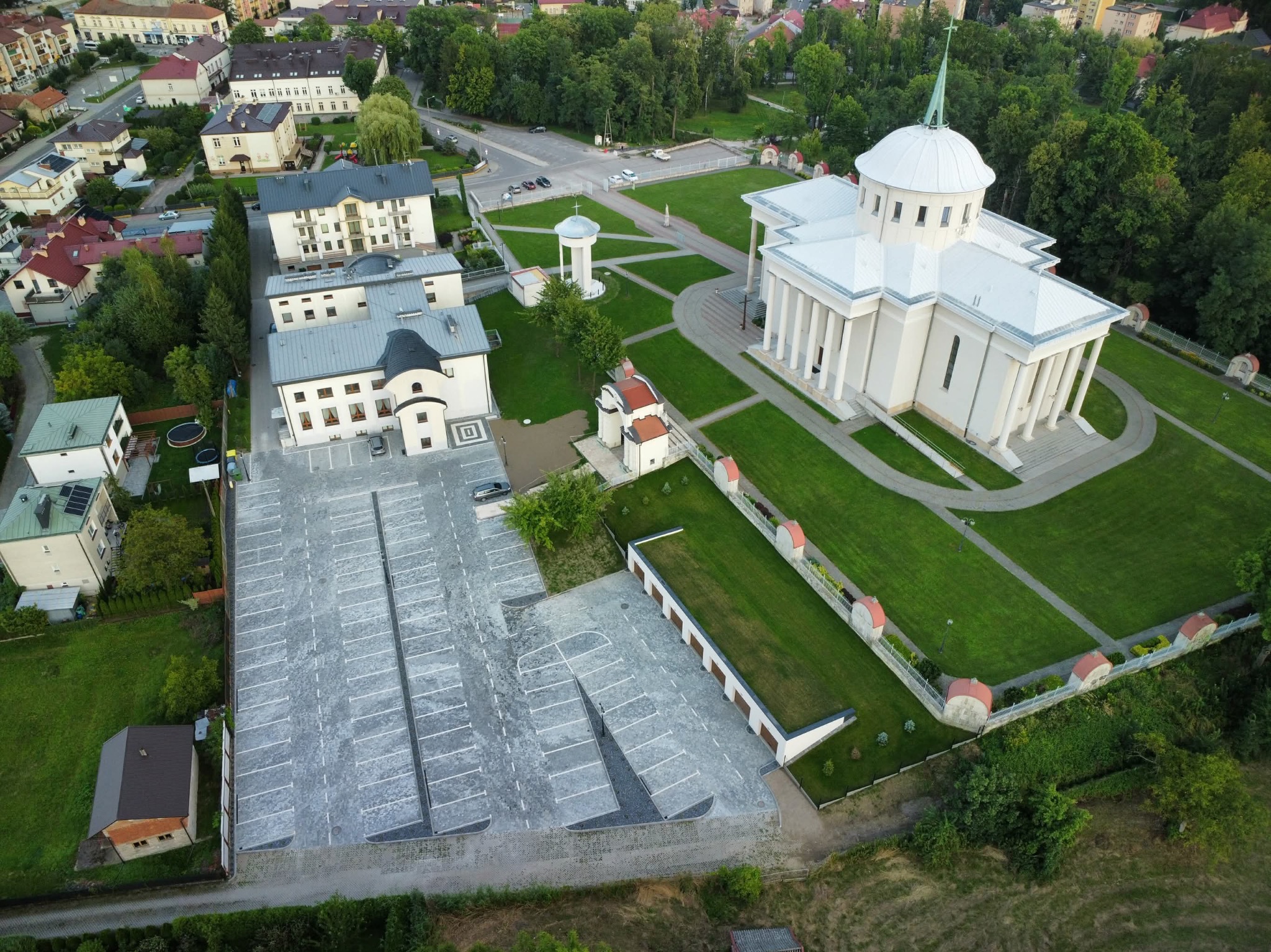 Widok z drona na duży parking z kostki brukowej obok białego kościoła z kolumnami, otoczonego zielenią i budynkami mieszkalnymi w słoneczny dzień.