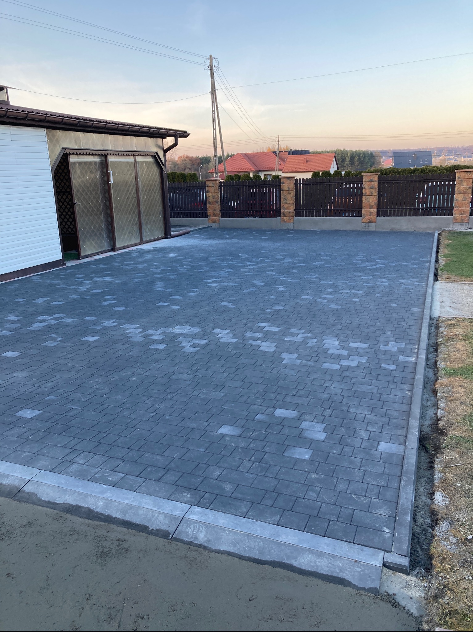 Ułożona, szara kostka brukowa na podjeździe, z widocznymi obrzeżami i fragmentem budynku z bramą garażową w tle. Regularny wzór kostki, równa powierzchnia.