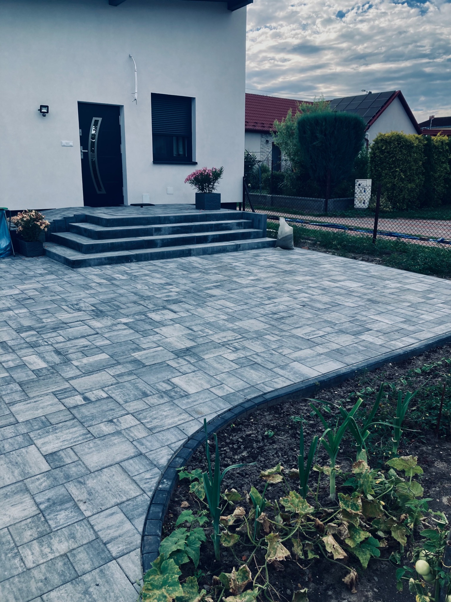 Szara kostka brukowa ułożona na podjeździe przed domem z czterema schodami wejściowymi, obrzeża z ciemnej kostki, rabata z warzywami przy krawędzi podjazdu, pochmurne niebo w tle.