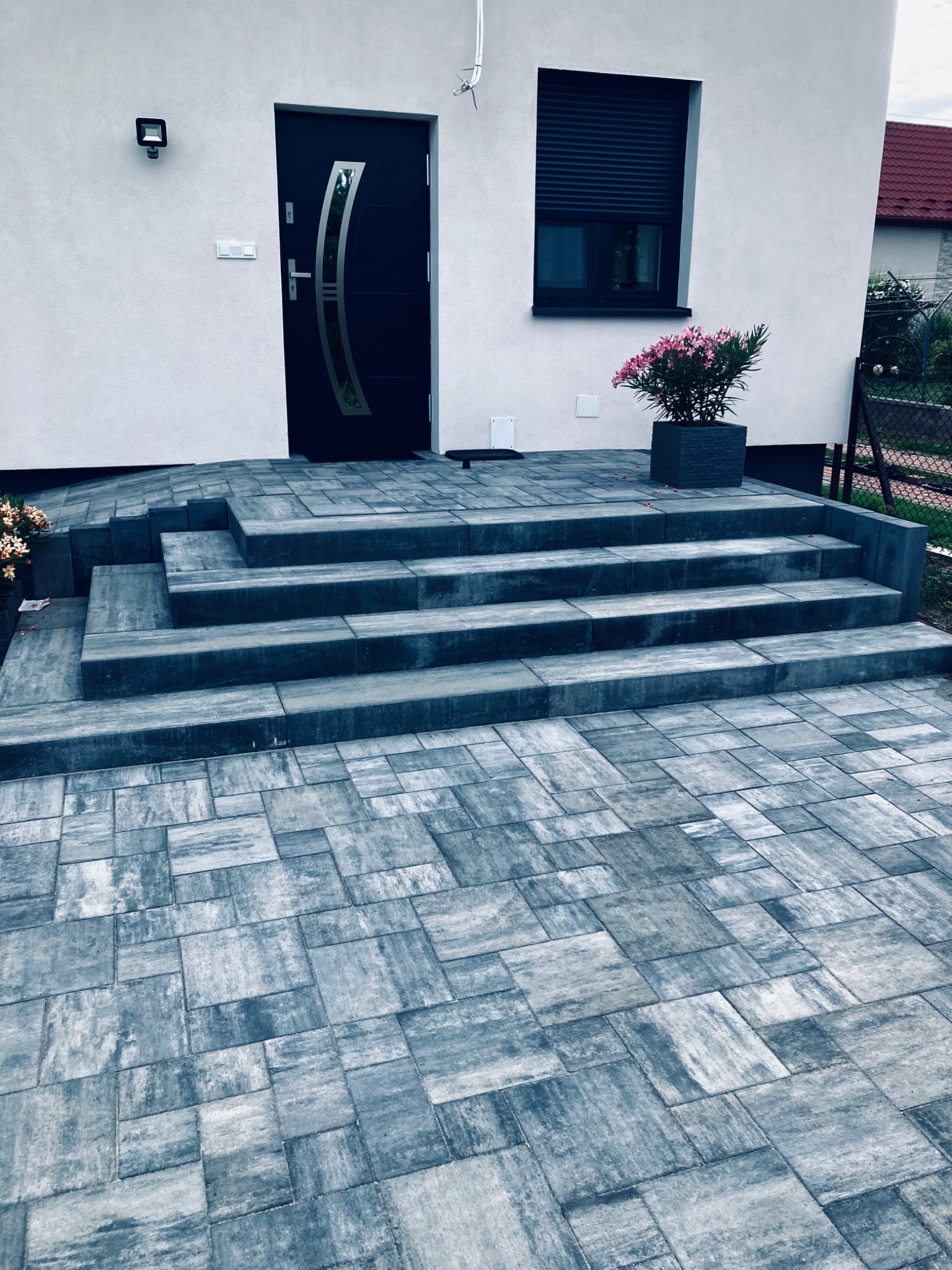 Ułożona kostka brukowa prowadząca do wejścia domu w Dąbrowie Tarnowskiej, z czterostopniowymi schodami i donicą z kwiatami obok. Kostka w odcieniach szarości, ułożona w nieregularny wzór.