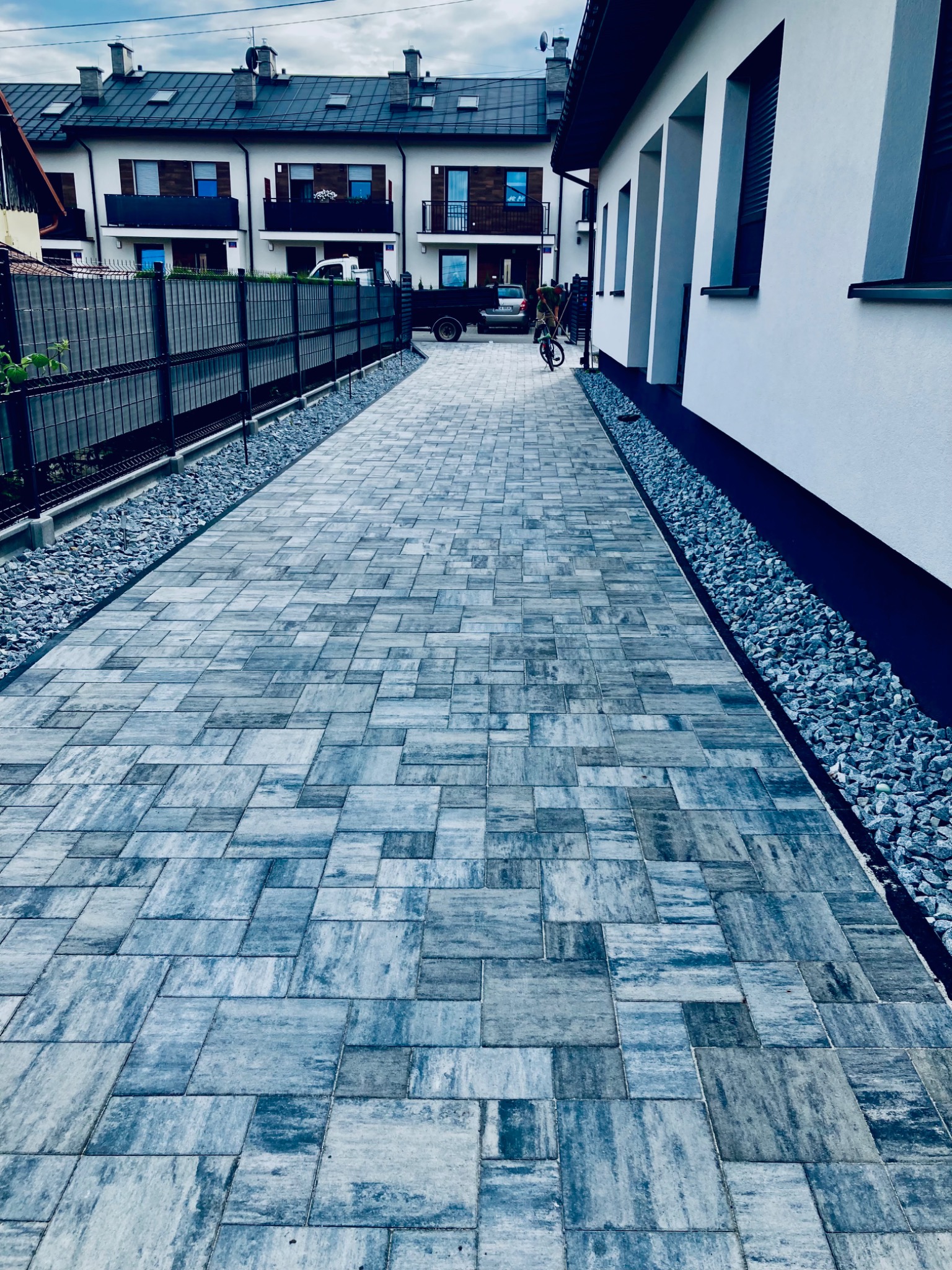 Nowo ułożona, szara kostka brukowa prowadząca wzdłuż białego budynku, oddzielona od niego pasem drobnych kamieni; w tle widoczne inne budynki mieszkalne i ogrodzenie.