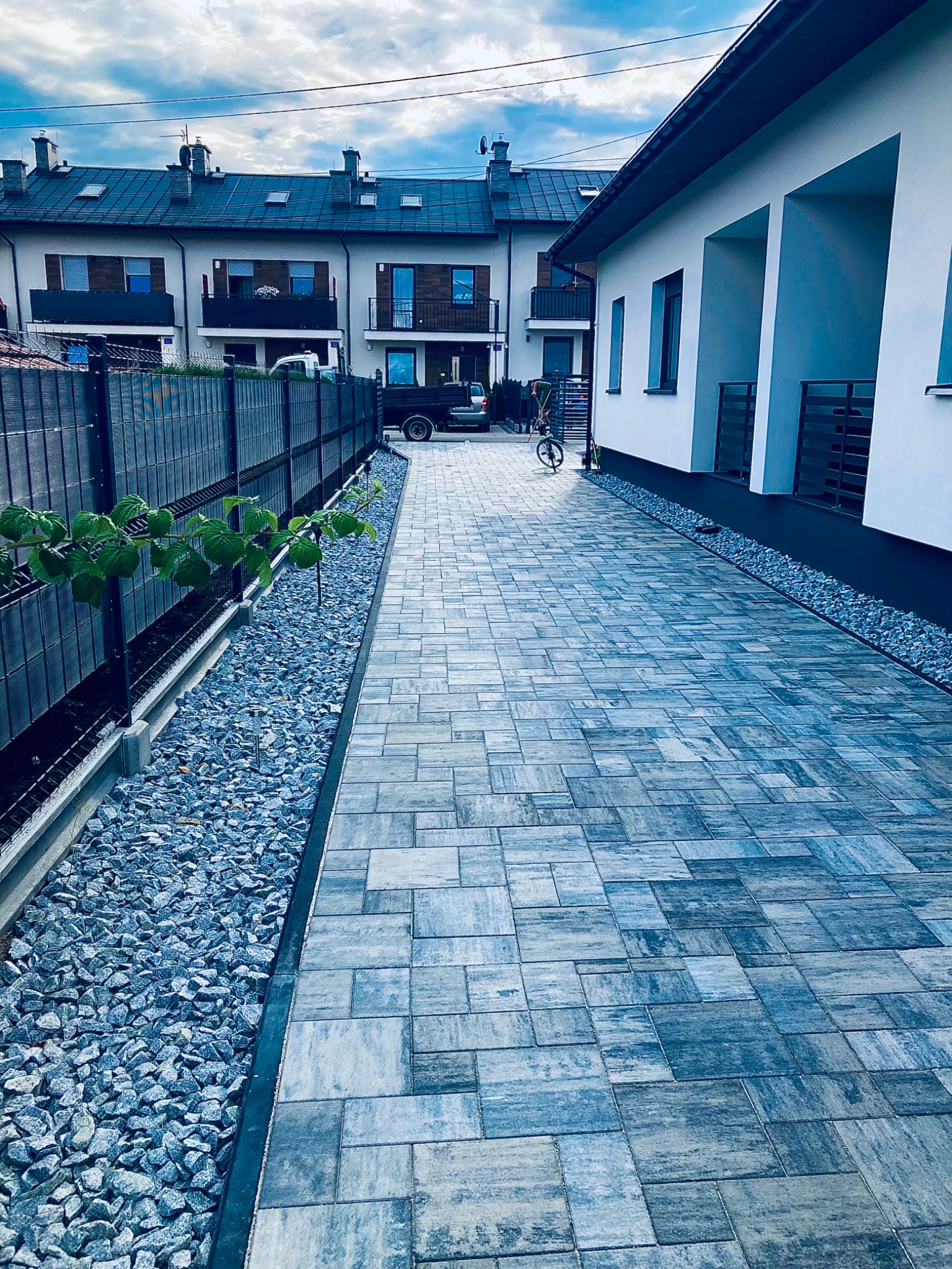 Ułożona kostka brukowa w odcieniach szarości prowadząca do nowoczesnego domu z białymi ścianami, obok czarny płot i pas grysu z niskimi roślinami.