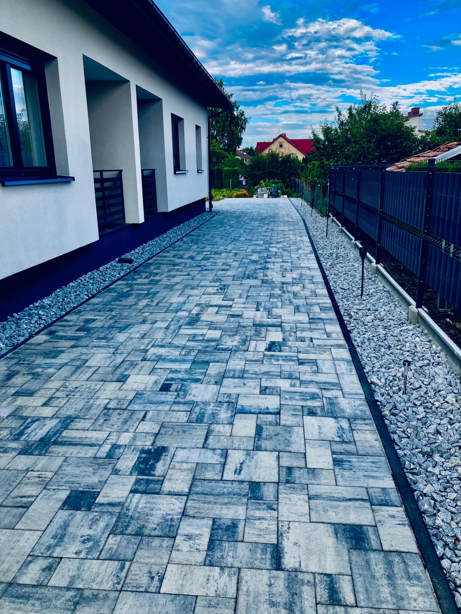 Ułożona nawierzchnia z szaro-niebieskiej kostki brukowej o nieregularnym wzorze, obok nowoczesny budynek z białymi ścianami i ciemnymi oknami, oddzielona od trawnika pasem drobnych kamieni