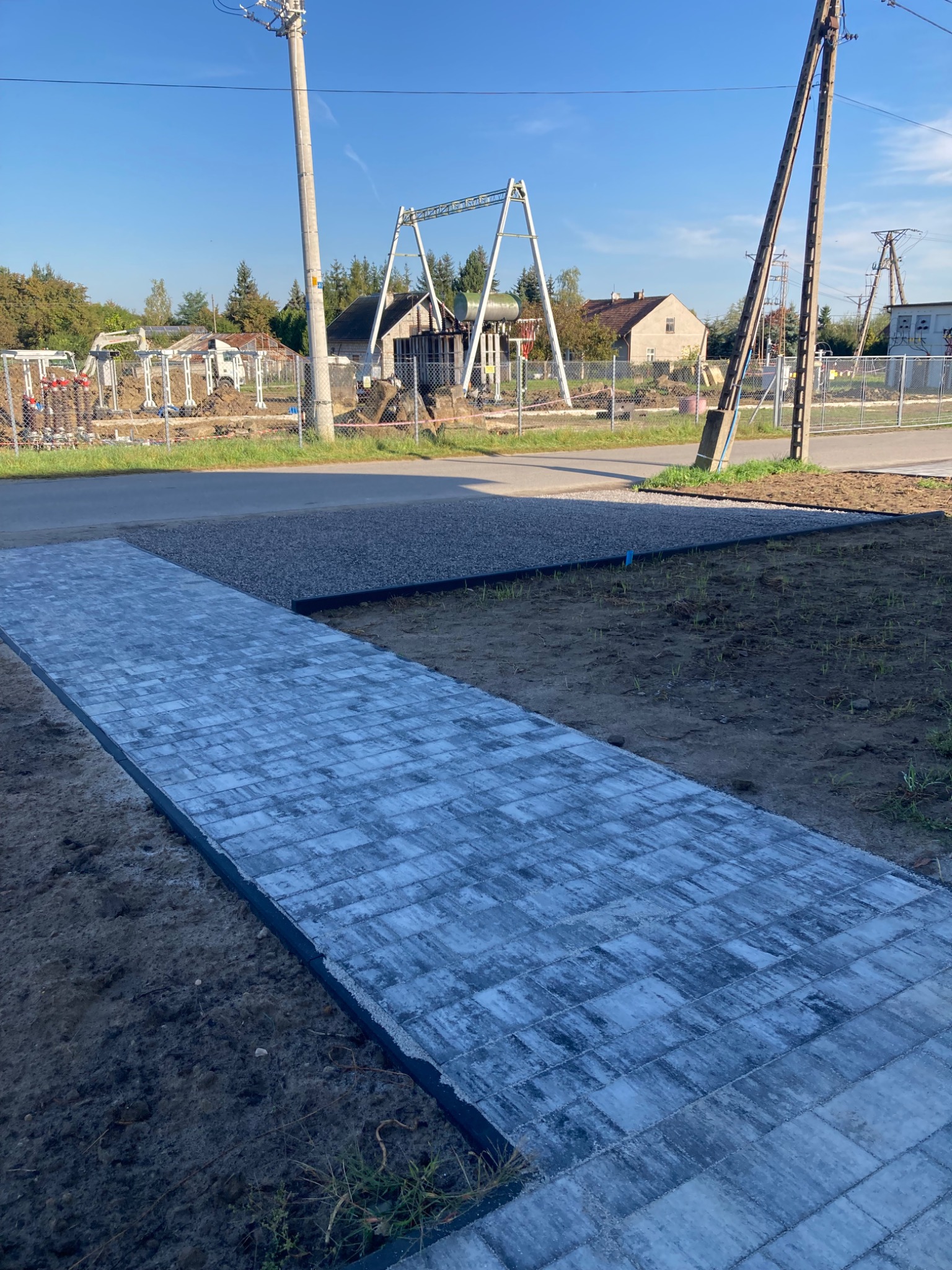 Nowo ułożony chodnik z szarych kostek brukowych prowadzący do obszaru z wysypanym żwirem, w tle widoczne prace ziemne i sprzęt budowlany oraz słupy wysokiego napięcia.