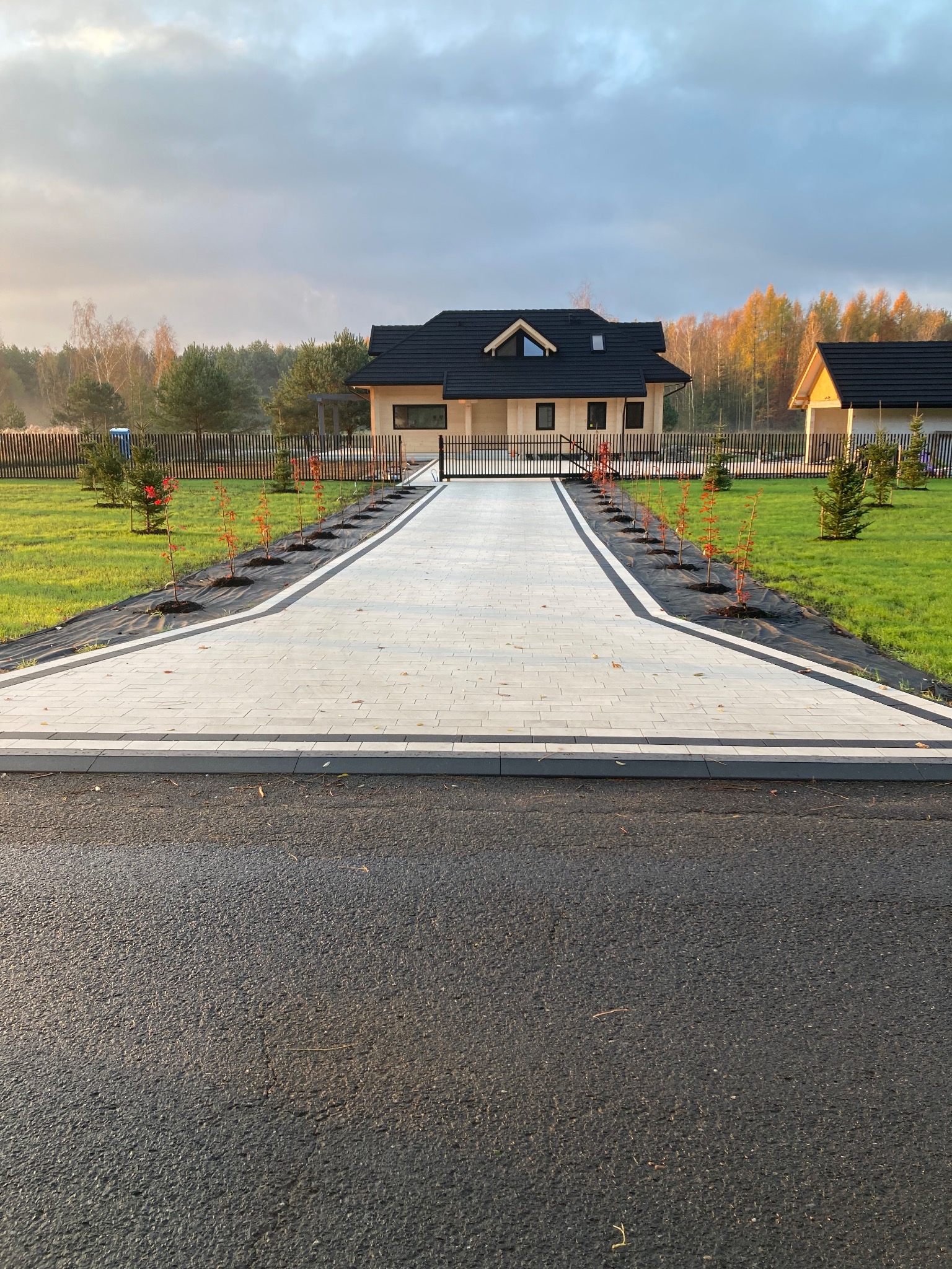 Nowoczesny podjazd z jasnoszarej kostki brukowej prowadzący do domu z czarnym dachem, obrzeża wykończone ciemniejszą kostką, po bokach posadzone młode drzewka.