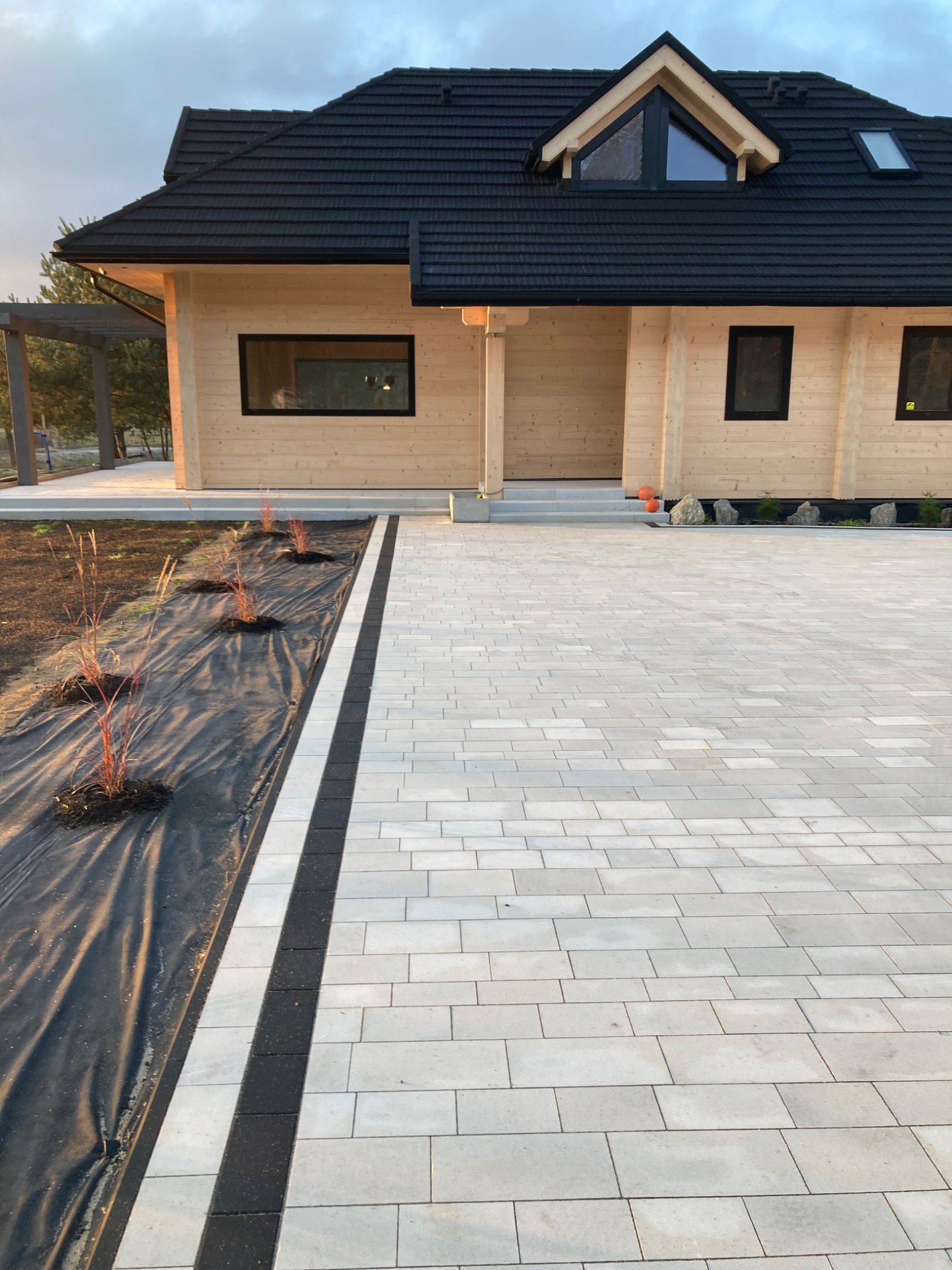 Nowoczesny dom z jasnego drewna z czarną dachówką i nowo ułożonym jasnoszarym brukiem z ciemnymi akcentami na podjeździe, obok rabata z czarną agrowłókniną i młodymi roślinami.