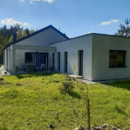 Nowoczesny dom jednorodzinny z płaskim dachem, wykończony jasnoszarym tynkiem, z widocznym tarasem i ogrodem w słoneczny dzień.