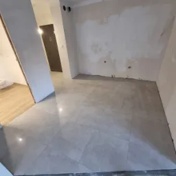 Ułożone szare płytki podłogowe w trakcie remontu mieszkania w Szczekocinach, widoczne niedoskonałości na ścianach przygotowanych do dalszych prac, w tle fragment łazienki z beżowymi płytkami.