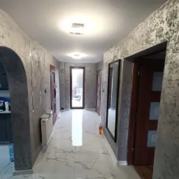 Korytarz w mieszkaniu po remoncie, ściany pokryte dekoracyjnym tynkiem w odcieniach szarości z efektem przetarć, biała marmurowa podłoga, widoczne drzwi i fragment zabudowy kuchennej.