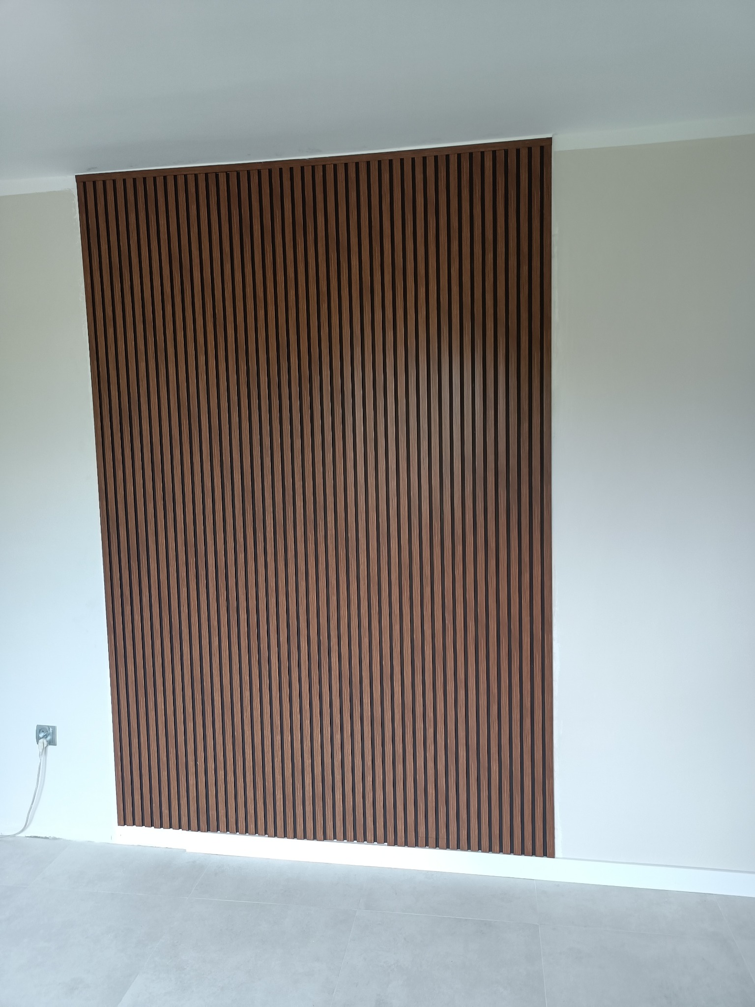 Panel ścienny z drewnianych listew w odcieniach brązu, zamontowany na białej ścianie w minimalistycznym wnętrzu z szarą podłogą.