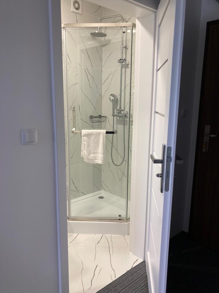 Nowoczesna kabina prysznicowa z białą ramą i szklanymi drzwiami, wykończona płytkami imitującymi marmur z czarnymi żyłami, widok z uchylonych białych drzwi łazienki.