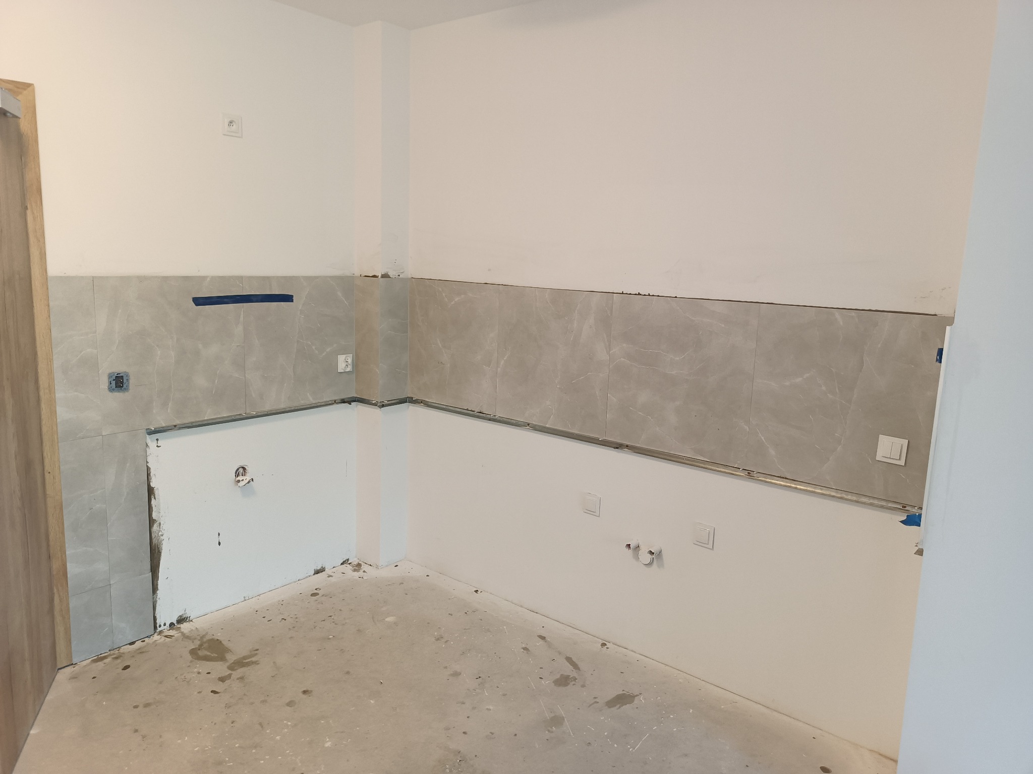 Wnętrze w trakcie prac przygotowawczych do montażu zabudowy kuchennej: fragment ściany z ułożonymi szarymi płytkami imitującymi marmur, widoczne wyprowadzenia instalacji elektrycznej i wodnej...