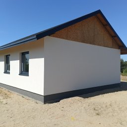 QBYG firma budowlana - Nowoczesny, parterowy dom z białą elewacją i grafitowymi oknami, widoczne wykończenie dachu i fragment ściany szczytowej z płyt OSB, otoczenie piaszczyste.