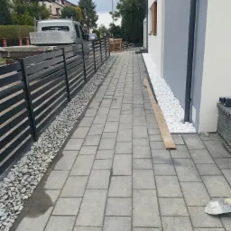 Ścieżka z szarej kostki brukowej obok budynku z szaro-białą elewacją, oddzielona od czarnego ogrodzenia pasem białych i szarych kamieni, widoczny samochód dostawczy w tle.