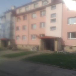 QBYG firma budowlana - Wielorodzinny budynek mieszkalny z elewacją w odcieniach różu i beżu, widziany z poziomu ulicy w słoneczny dzień, z widocznymi elementami ocieplenia przy gruncie.