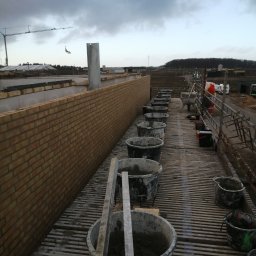 QBYG firma budowlana - Wznoszenie muru z żółtej cegły na rusztowaniu, widoczne wiadra z zaprawą, w tle krajobraz budowy pod pochmurnym niebem.