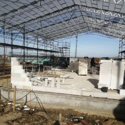 QBYG firma budowlana - Wznoszenie muru z bloczków na fundamencie wewnątrz konstrukcji stalowej z zadaszeniem, widoczne rusztowania i sprzęt budowlany w tle.