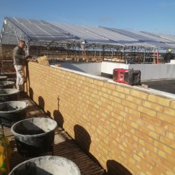 QBYG firma budowlana - Murarz w trakcie wznoszenia muru z żółtej cegły na zewnątrz budynku, widoczne wiadra z zaprawą i konstrukcja zadaszenia w tle.