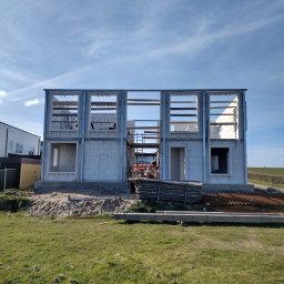 QBYG firma budowlana - Surowy stan dwukondygnacyjnego domu z białych bloczków, widoczne metalowe ramy okienne i rusztowanie wewnątrz, na pierwszym planie trawa i materiały budowlane.