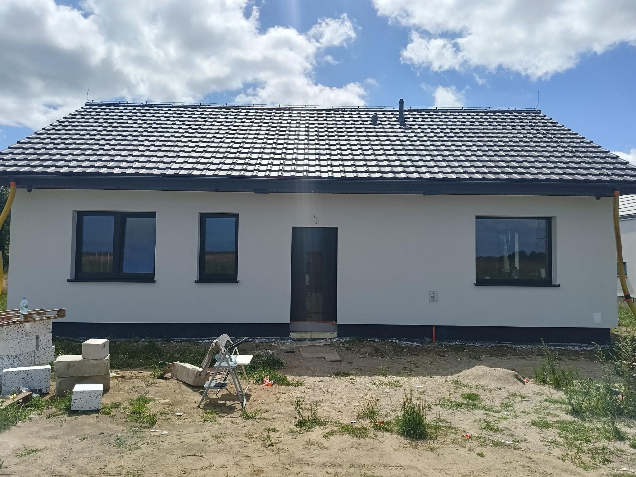 Otynkowany, parterowy dom jednorodzinny z czarną dachówką i stolarką okienną, widok z przodu, z widocznymi materiałami budowlanymi i drabinką na posesji.