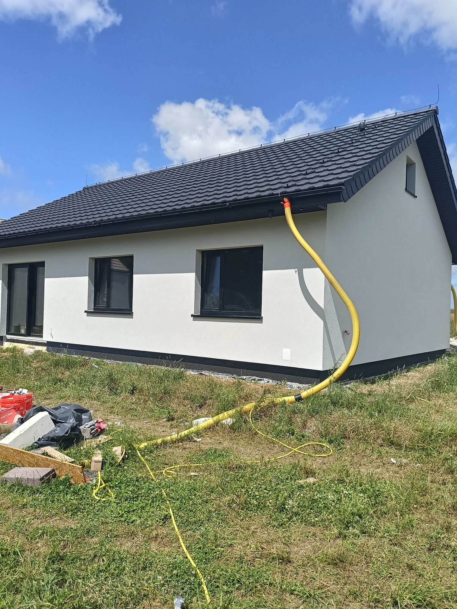 Ocieplony dom jednorodzinny z grafitową dachówką, widoczna żółta rura wentylacyjna wychodząca spod dachu i rozciągająca się na trawniku, oraz materiały budowlane w tle.