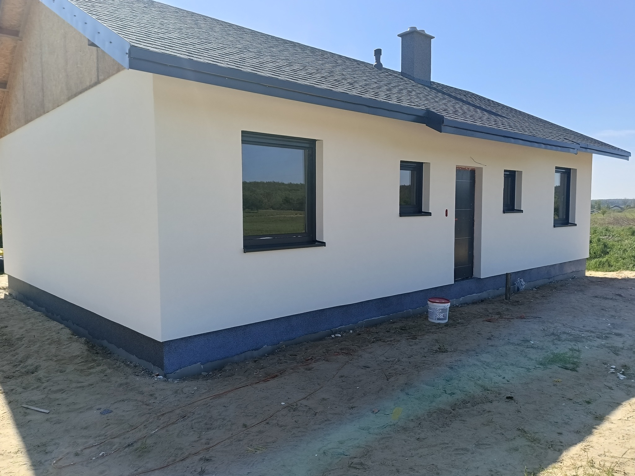 Nowo otynkowany, parterowy dom jednorodzinny z szarą podmurówką i grafitową stolarką okienną, widok z zewnątrz, w tle błękitne niebo.