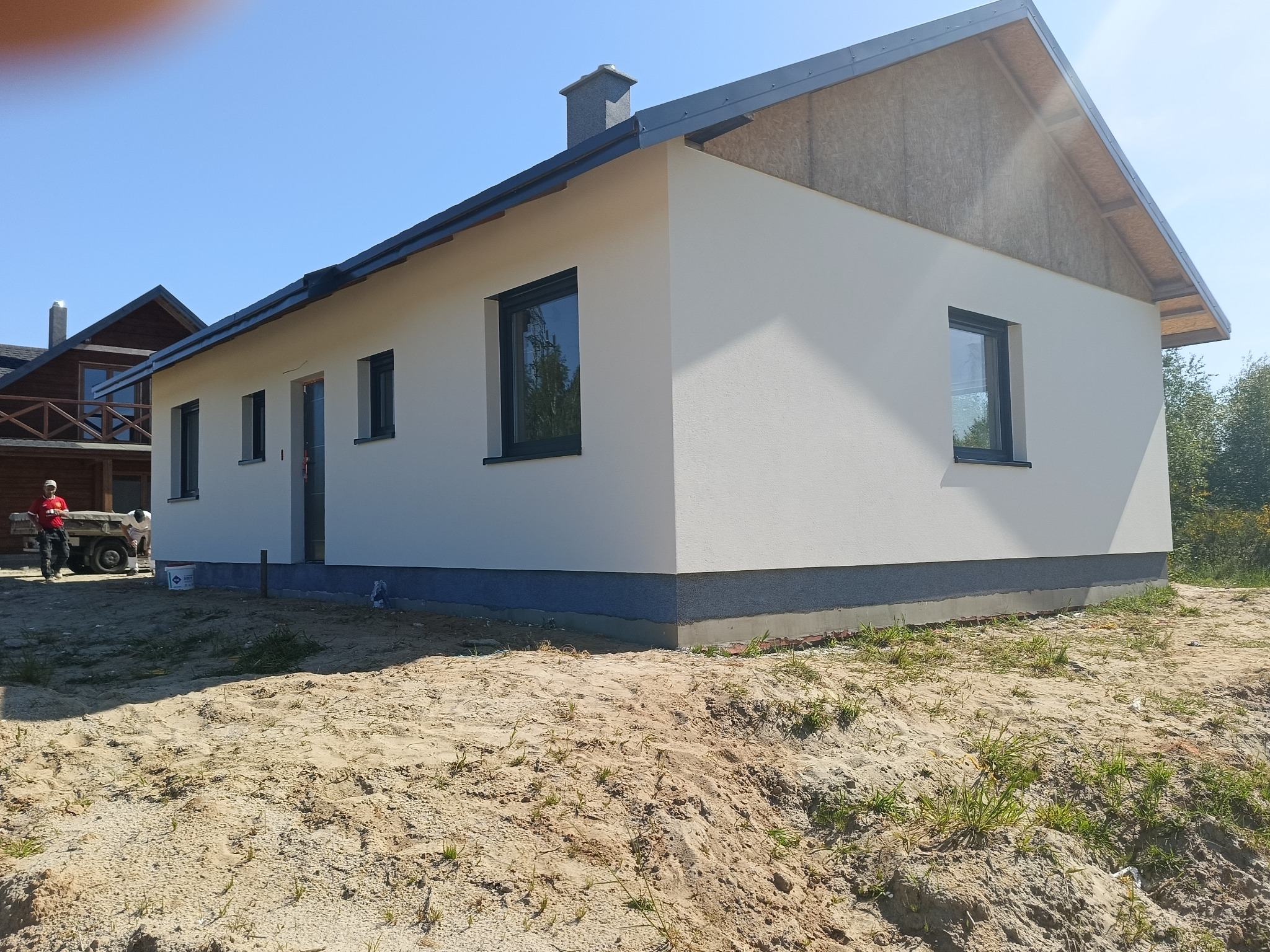 Nowo otynkowany, parterowy dom z szarą podmurówką i ciemnymi oknami, widoczny fragment niezabudowanej połaci dachu i ekipa budowlana z taczką w tle.