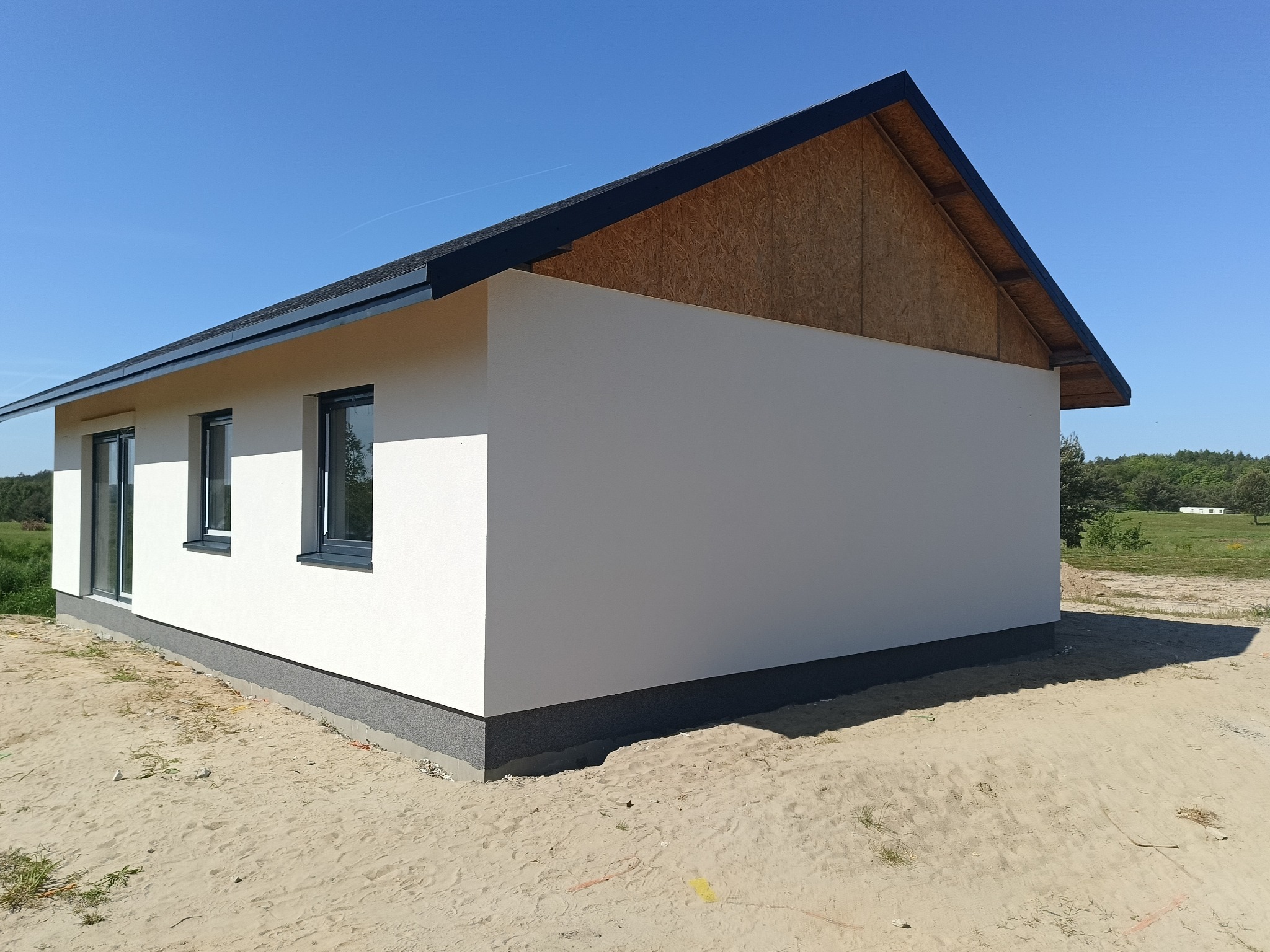 Nowoczesny, parterowy dom z białą elewacją i grafitowymi oknami, widoczne wykończenie dachu i fragment ściany szczytowej z płyt OSB, otoczenie piaszczyste.
