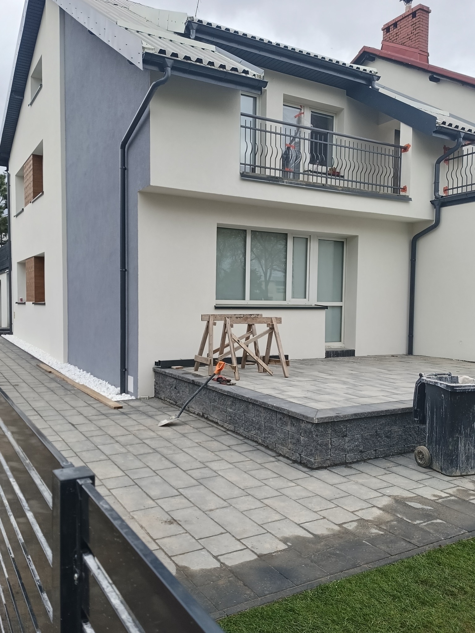 Dwukondygnacyjny dom jednorodzinny z szarą i białą elewacją, widoczne rusztowanie, narzędzia i kostka brukowa na podwórku, sugerujące prace wykończeniowe.