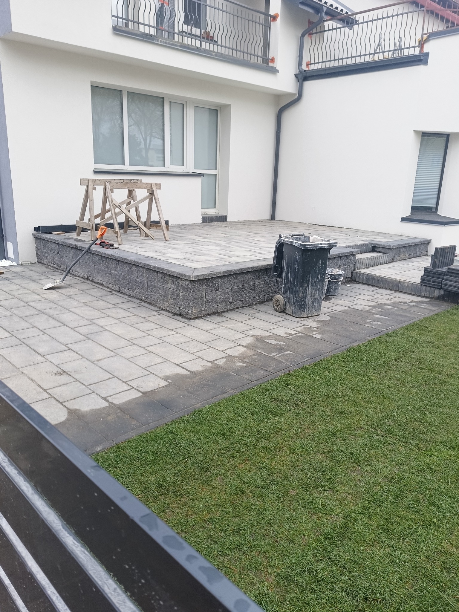 Nowoczesny taras z szarej kostki brukowej przy białym domu, widoczne narzędzia i resztki cementu, sugerujące trwające prace wykończeniowe.