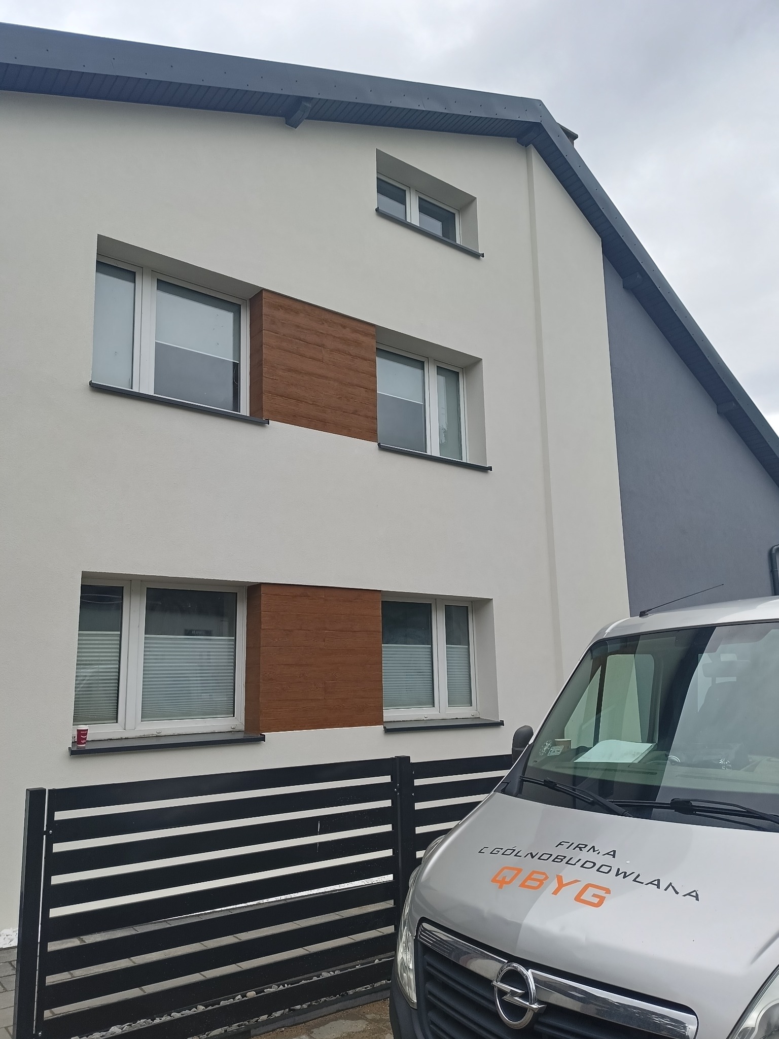 Nowoczesna elewacja domu z białym tynkiem i dekoracyjnymi panelami drewnopodobnymi, widoczny fragment firmowego samochodu dostawczego z logo firmy budowlanej.