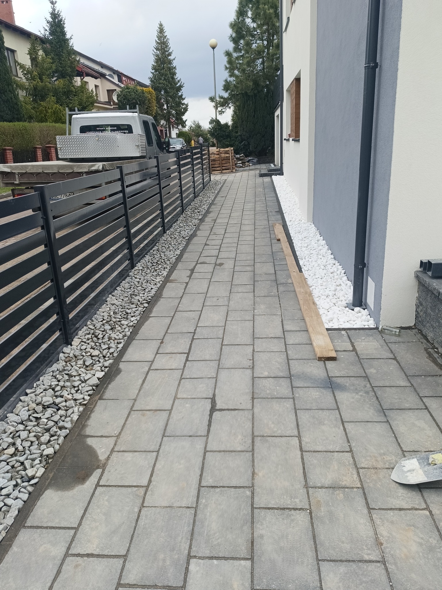 Ścieżka z szarej kostki brukowej obok budynku z szaro-białą elewacją, oddzielona od czarnego ogrodzenia pasem białych i szarych kamieni, widoczny samochód dostawczy w tle.