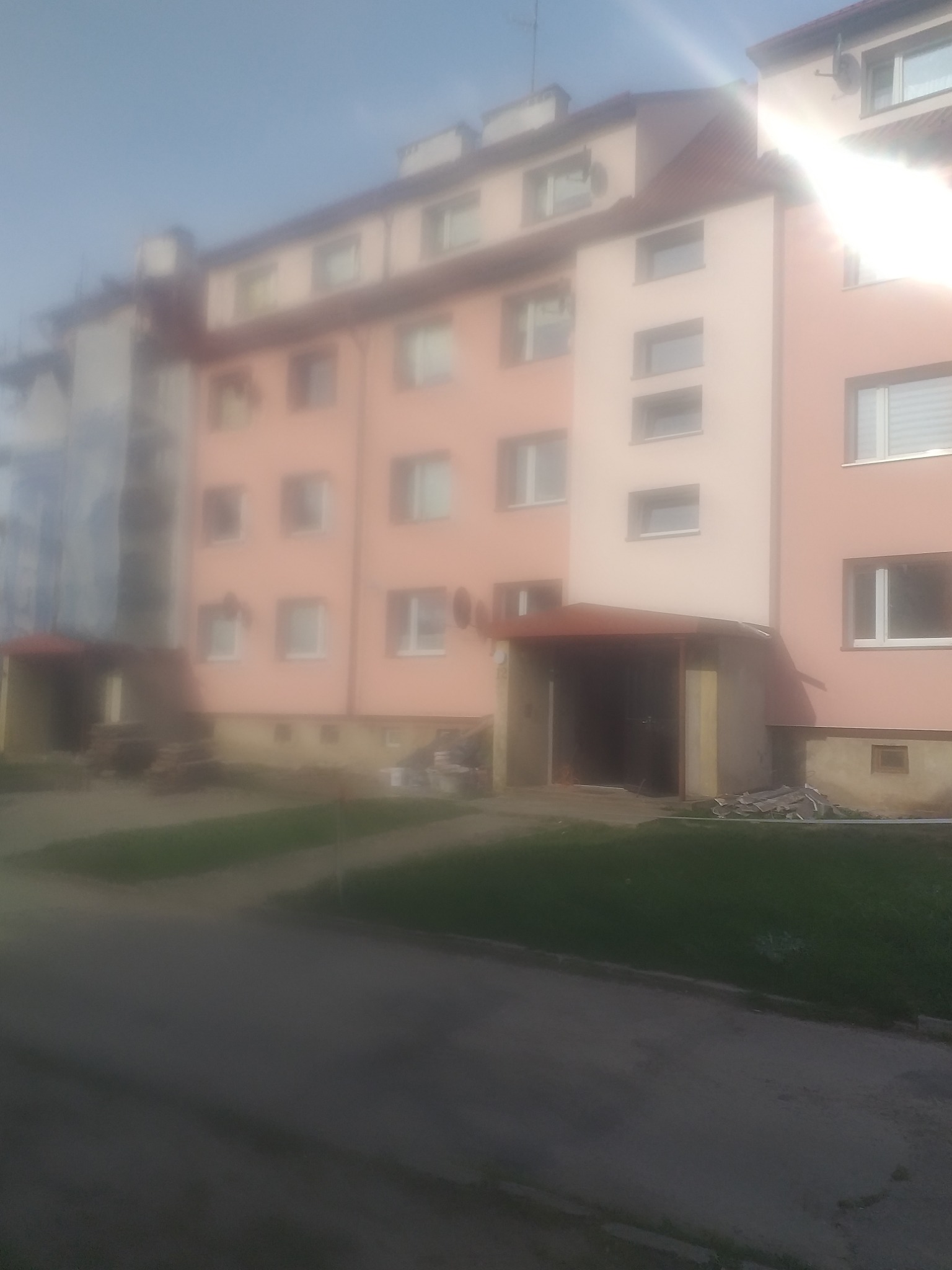 Wielorodzinny budynek mieszkalny z elewacją w odcieniach różu i beżu, widziany z poziomu ulicy w słoneczny dzień, z widocznymi elementami ocieplenia przy gruncie.