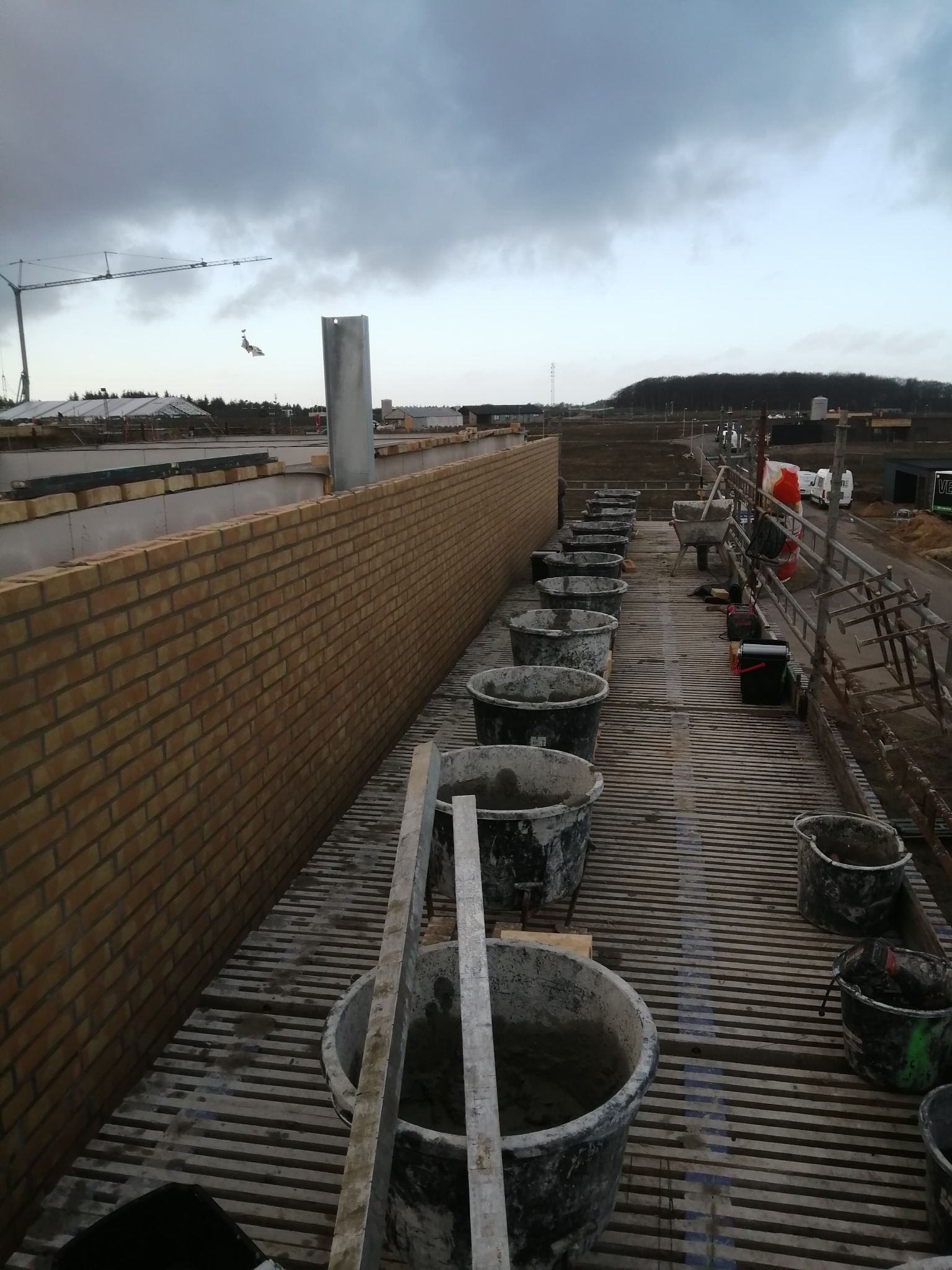 Wznoszenie muru z żółtej cegły na rusztowaniu, widoczne wiadra z zaprawą, w tle krajobraz budowy pod pochmurnym niebem.