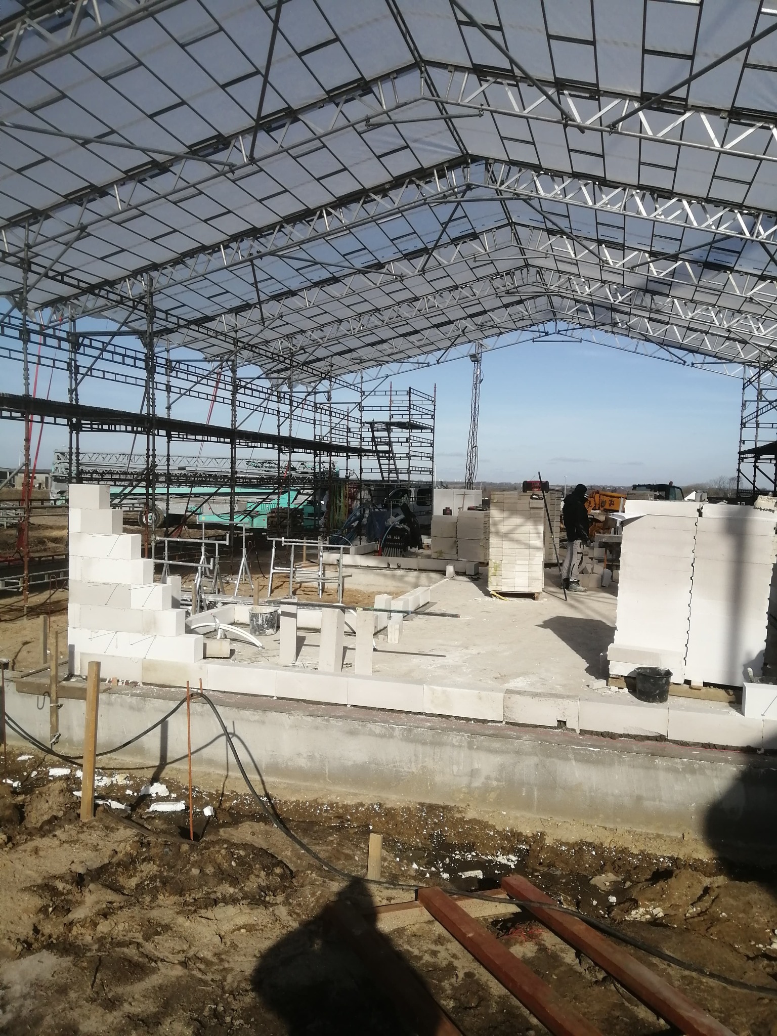 Wznoszenie muru z bloczków na fundamencie wewnątrz konstrukcji stalowej z zadaszeniem, widoczne rusztowania i sprzęt budowlany w tle.