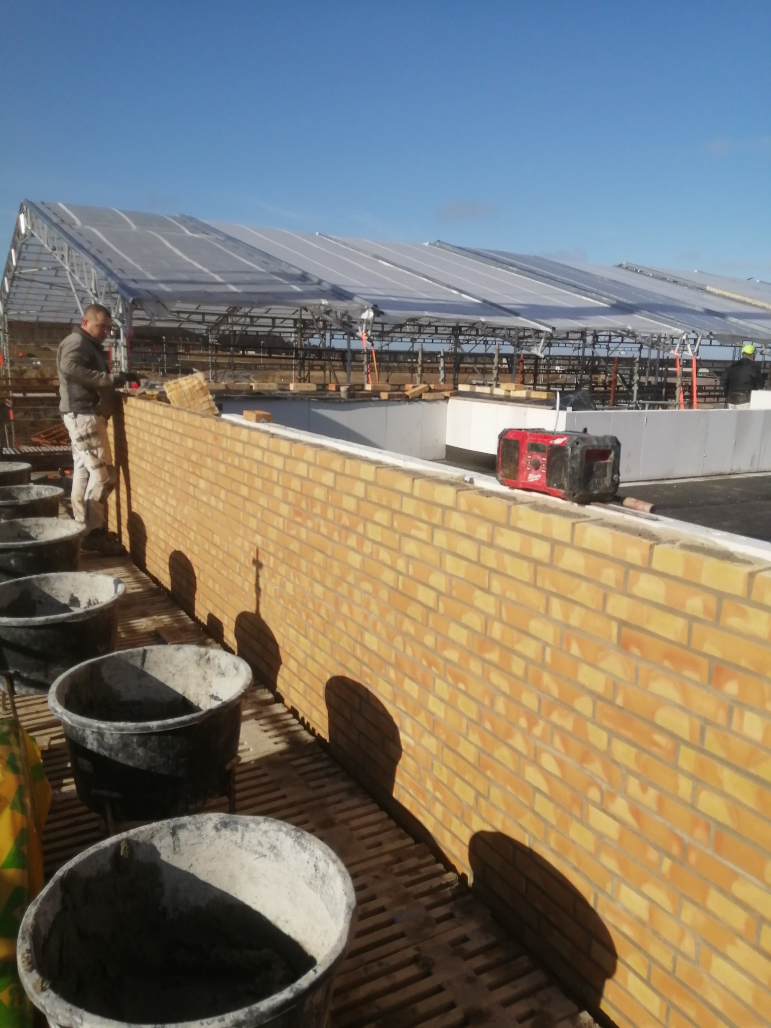 Murarz w trakcie wznoszenia muru z żółtej cegły na zewnątrz budynku, widoczne wiadra z zaprawą i konstrukcja zadaszenia w tle.