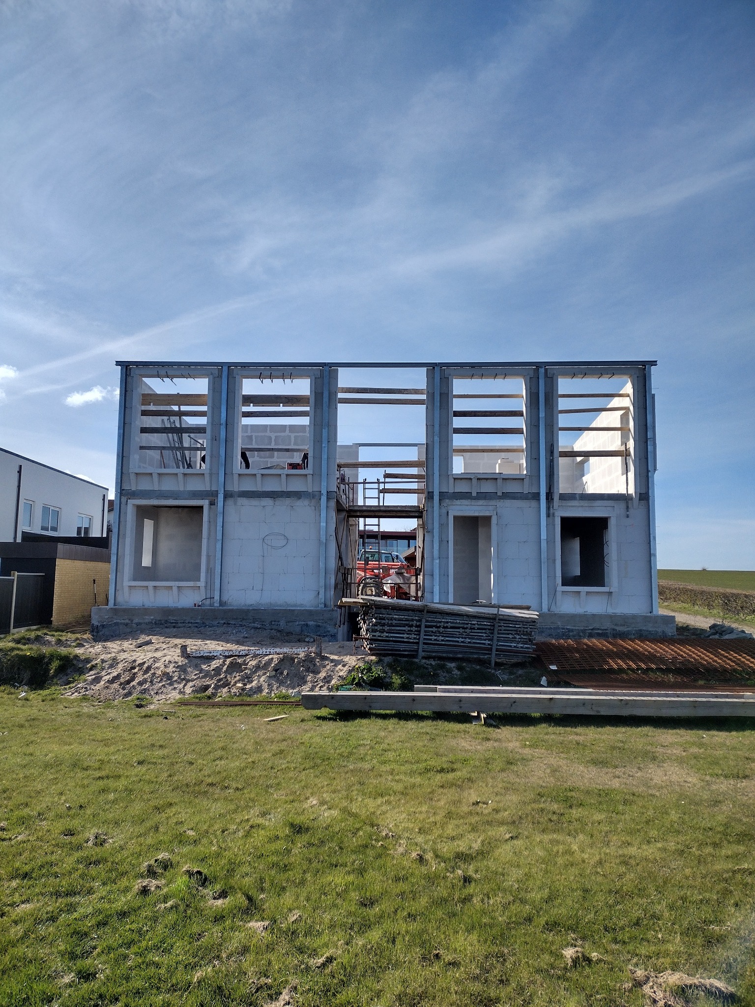 Surowy stan dwukondygnacyjnego domu z białych bloczków, widoczne metalowe ramy okienne i rusztowanie wewnątrz, na pierwszym planie trawa i materiały budowlane.