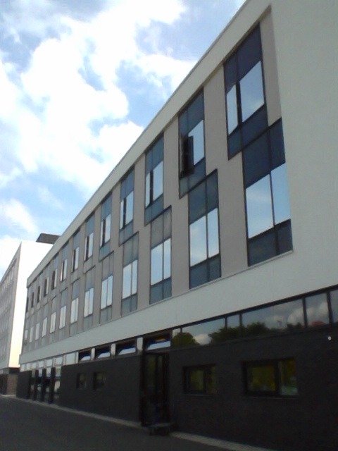 Nowoczesny budynek biurowy z szarą elewacją i ciemnymi ramami okiennymi, widok z dołu na tle pochmurnego nieba, jedno okno otwarte.