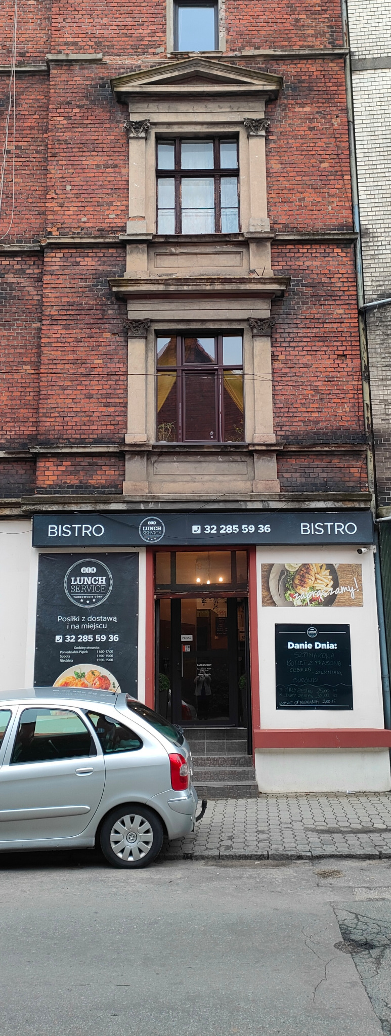 Fasada budynku z szyldem Bistro Lunch Service, menu dnia na tablicy, srebrny samochód częściowo zasłaniający wejście.