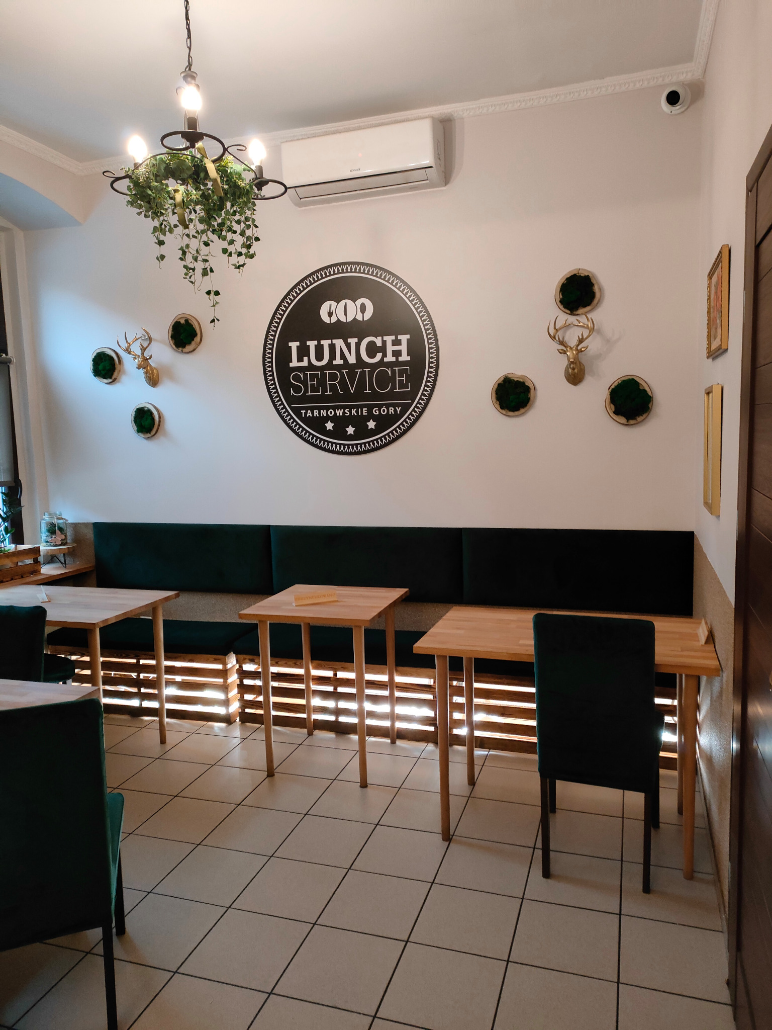 Jasne wnętrze lokalu gastronomicznego z drewnianymi stolikami, zielonymi krzesłami i sofą wzdłuż ściany, ozdobionej logo 'Lunch Service Tarnowskie Góry', sztucznymi porożami jelenia i roślinami...