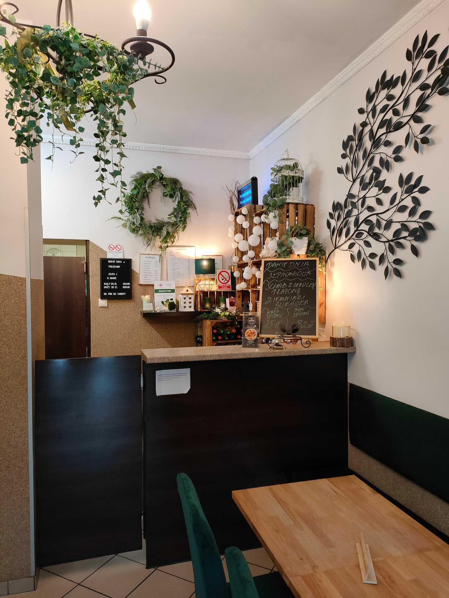 Wystrój wnętrza lokalu gastronomicznego z elementami roślinnymi: wisząca zieleń nad ladą, wieniec, dekoracja ścienna w kształcie drzewa i tablica z menu.