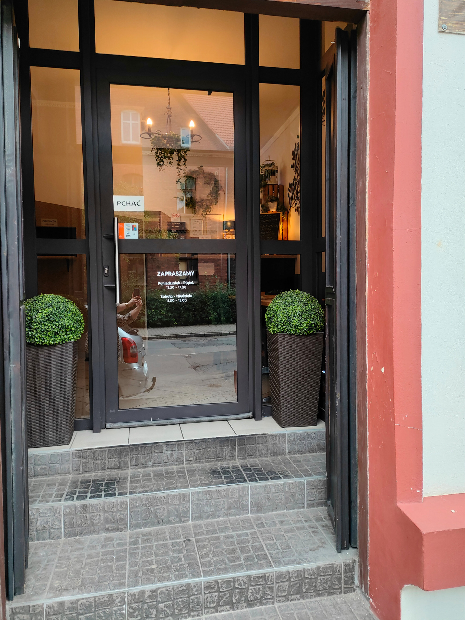 Wejście do lokalu z przeszklonymi drzwiami, na których widnieje informacja o godzinach otwarcia oraz logo firmy PCHAĆ. Po bokach drzwi stoją donice z zielonymi kulistymi krzewami.