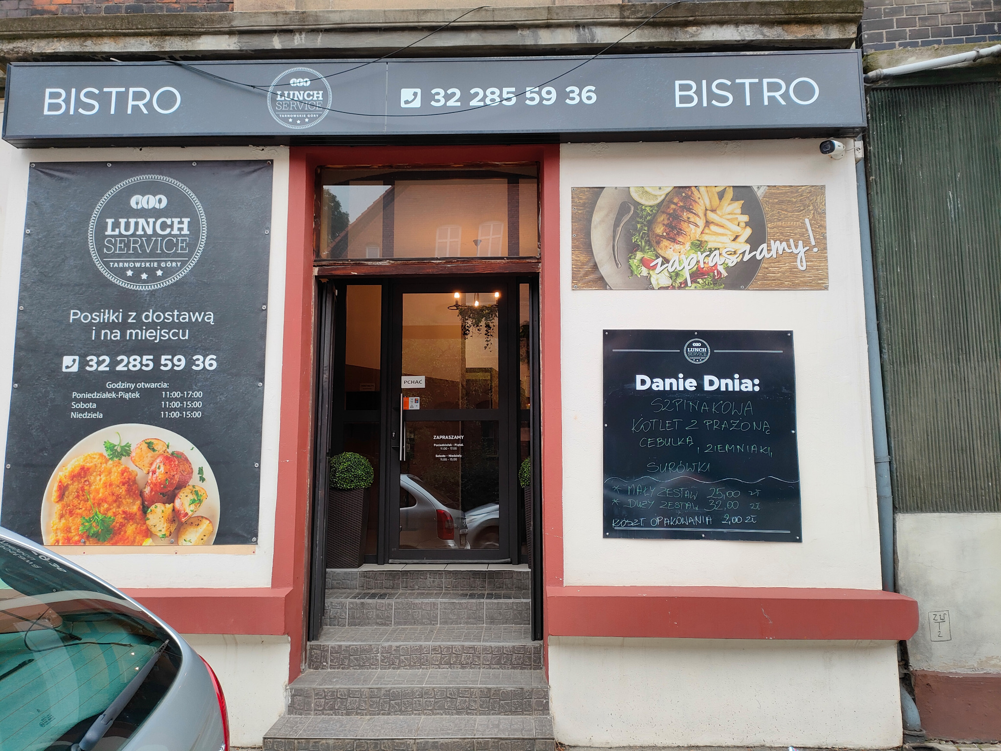 Fasada bistro z szyldem Lunch Service, menu dnia wypisane kredą na tablicy, informacja o dostawie i godzinach otwarcia, widok z zewnątrz, fragment samochodu w kadrze.