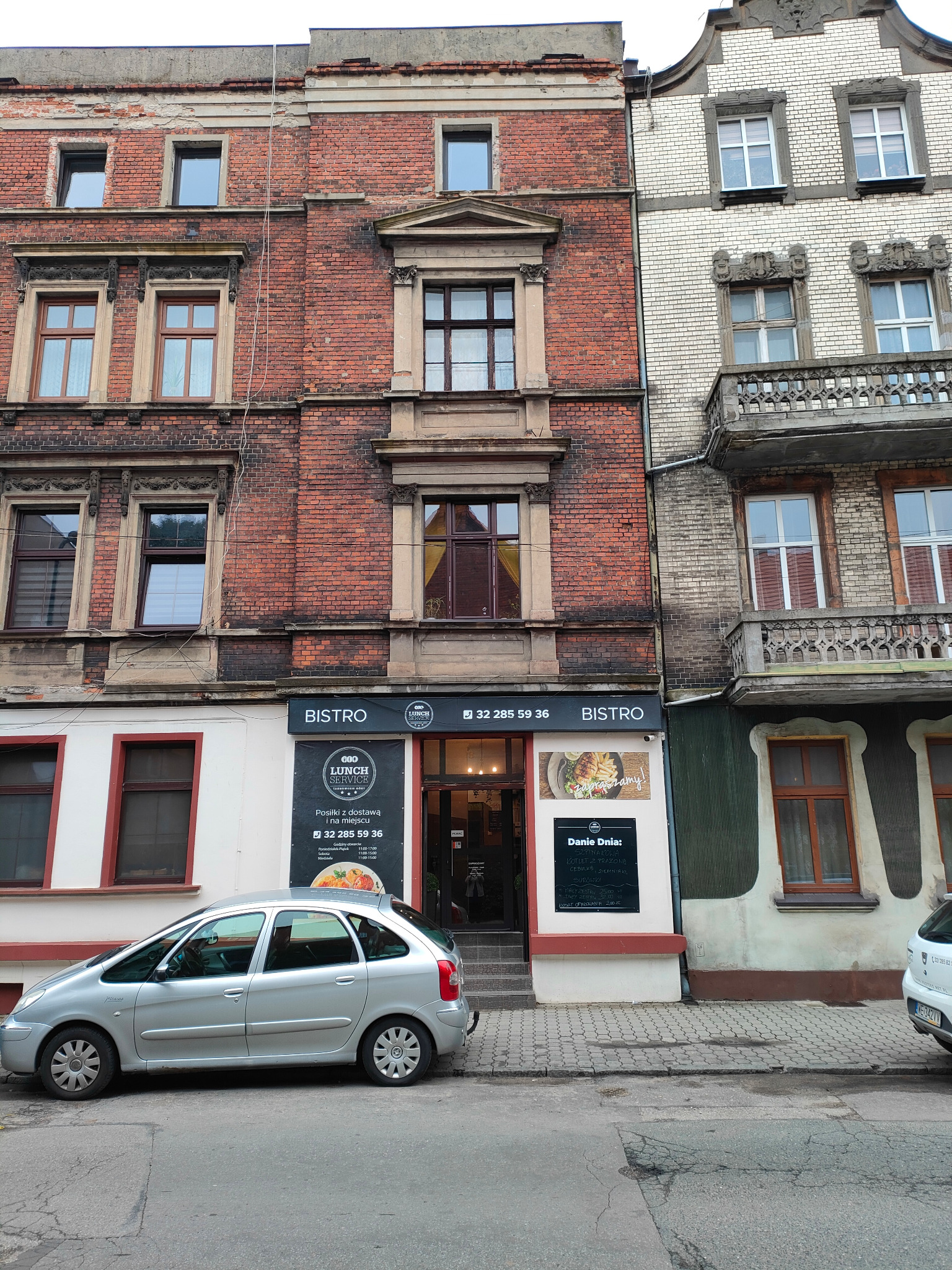 Fasada kamienicy z szyldem 'Bistro' i reklamą 'Lunch Service' oraz zaparkowanym srebrnym Citroenem Picasso przed wejściem.