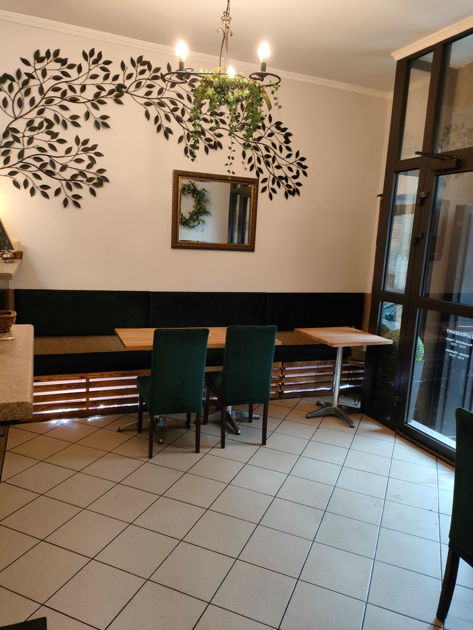Wnętrze restauracji z drewnianymi stołami, zielonymi krzesłami i dekoracją ścienną z motywem roślinnym, lustro z wieńcem na ścianie, widok na drzwi wejściowe.