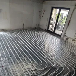 Wnętrze w trakcie instalacji ogrzewania podłogowego: białe rury ułożone na folii z czarną siatką, widoczne wyprowadzenia rur ze ściany, surowy stan wykończenia pomieszczenia, okno balkonowe w tle.