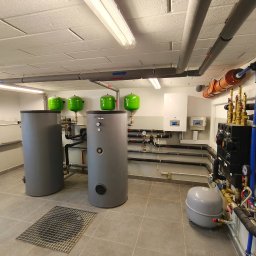 PROTERM Sebastian Jankowski - Kompletna kotłownia z dwoma szarymi zbiornikami, zielonymi naczyniami przeponowymi i licznymi rurami. Widoczne elementy sterujące i zabezpieczające instalację grzewczą.
