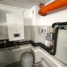 PROTERM Sebastian Jankowski - Nowoczesny, biały piec gazowy wiszący na ścianie, z podłączonymi rurami w izolacji, szary zbiornik wyrównawczy na podłodze, instalacja w pomieszczeniu technicznym.