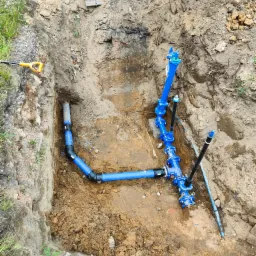 Niebieskie rury wodociągowe w wykopie ziemnym, widoczny fragment koparki i łopata.